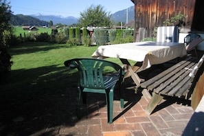 Een terras met een tafel en stoelen, een houten bankje, en uitzicht op een groen veld en bergen op de achtergrond.