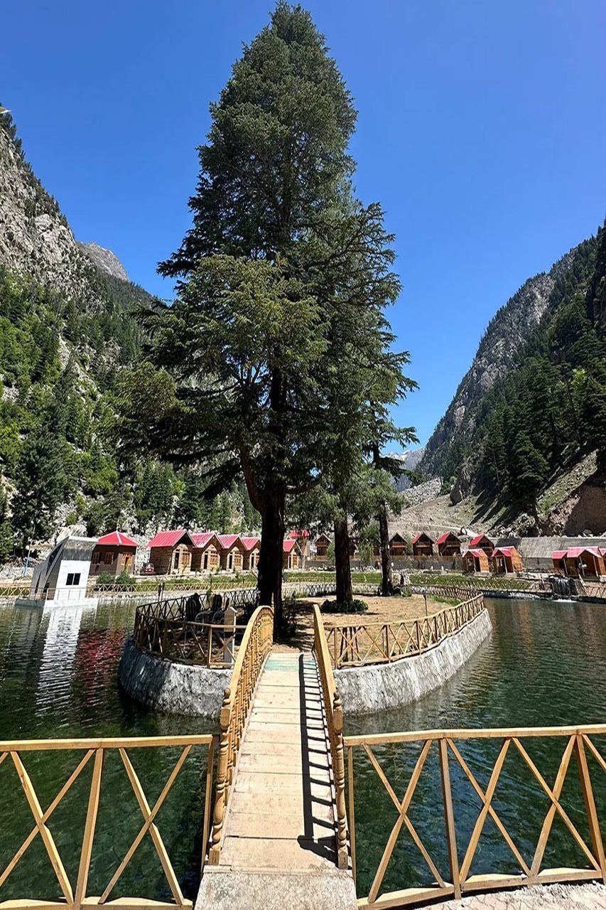 Un puente de madera que conduce a una hilera de cabañas con tejados rojos, rodeado de un paisaje montañoso y una masa de agua.