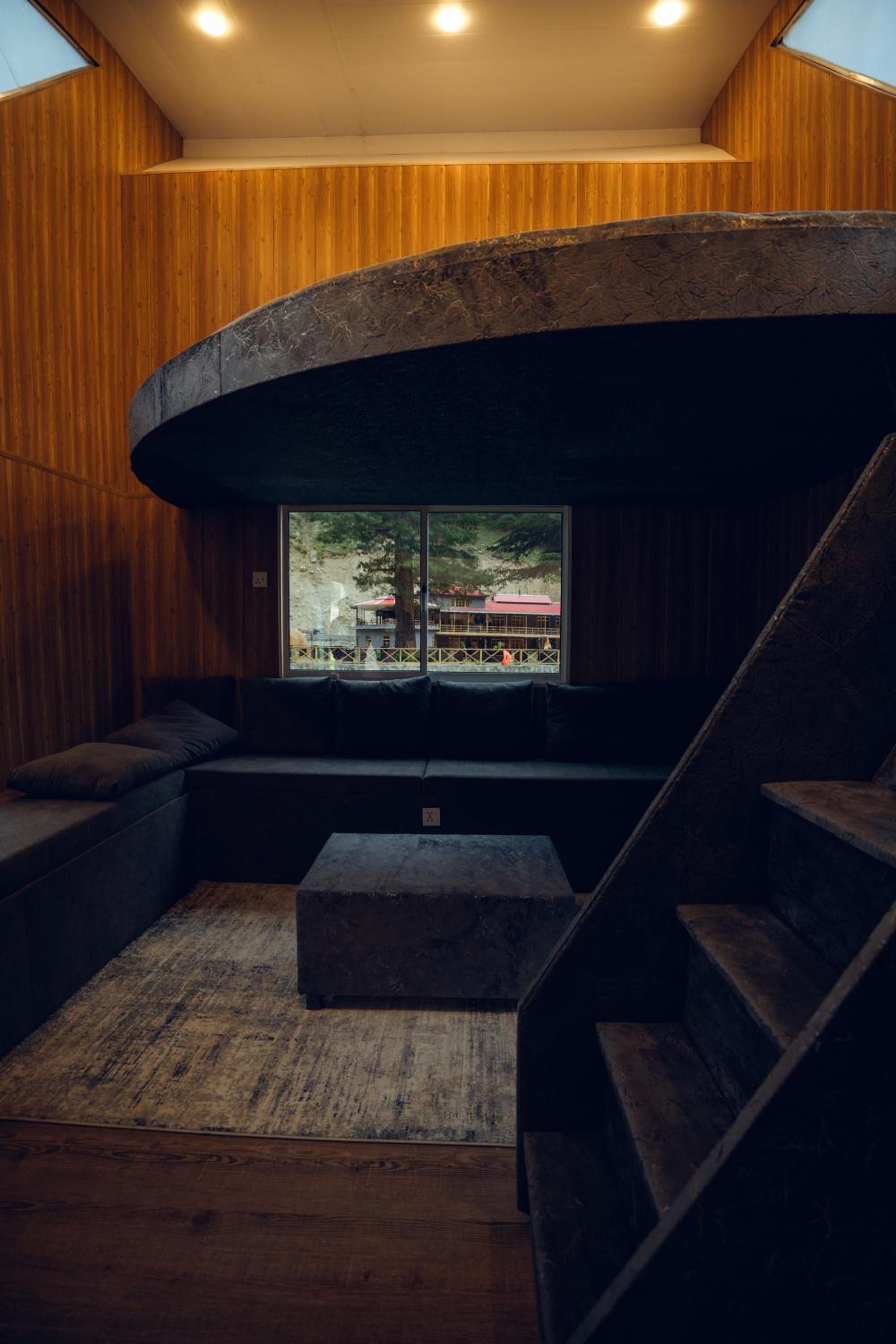 Un intérieur moderne avec une structure en pierre aux lignes courbes, des murs en bois et un espace salon offrant une vue sur un bâtiment à l’extérieur.