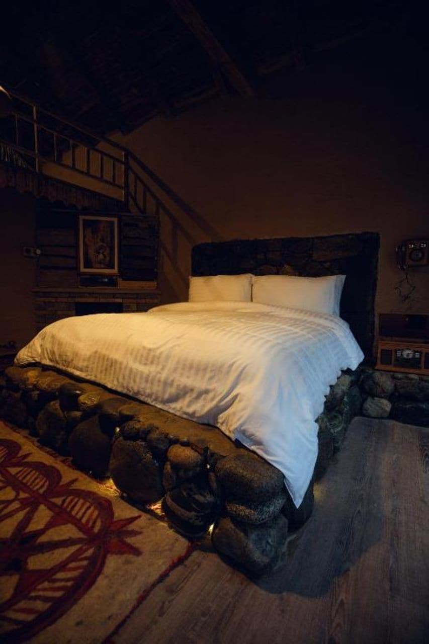 Una cama con ropa blanca, una pared de piedra y un cabecero de madera.