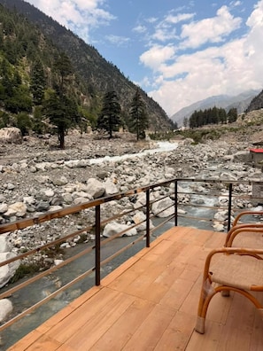 Una terraza de madera con barandilla, con vista a un lecho rocoso de río y montañas.