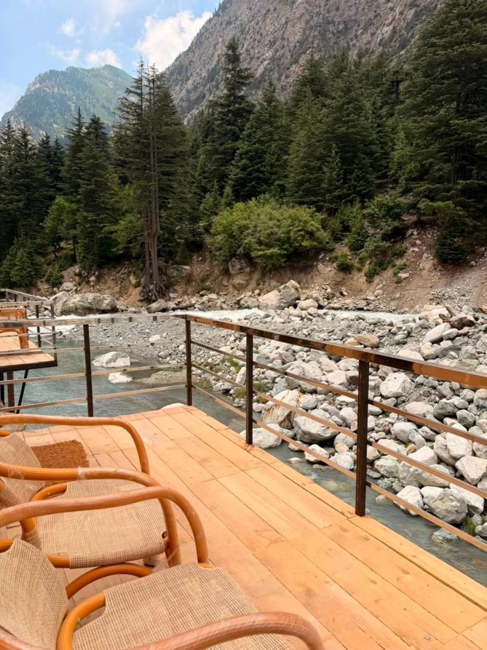 A wooden deck with chairs overlooking a rocky riverbed, surrounded by dense forest and mountains.