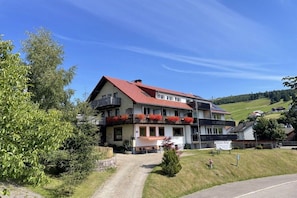 A two-story building with a red roof, balconies with red flowers, and a paved driveway leading to the entrance. - Holiday apartment with 2 bedrooms (Todtnauberg)