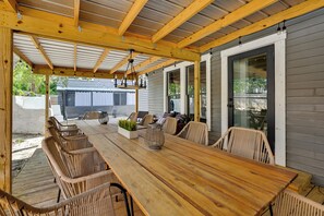 Une terrasse extérieure avec une table en bois et des chaises en osier, un luminaire suspendu et une vue sur une maison en arrière-plan.