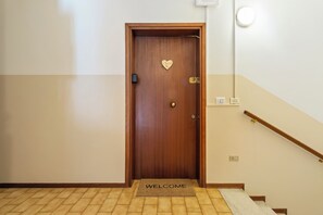 Een houten deur met een hartvormig plaquette, een deurmat met de tekst 'WELCOME' en een leuning op een trap.