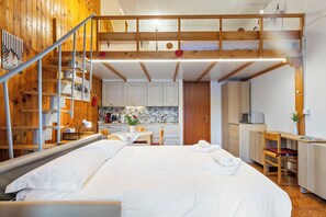 Een slaapkamer met een groot bed, een bureau met stoel, een keukenhoek en een zolderruimte met houten balken.