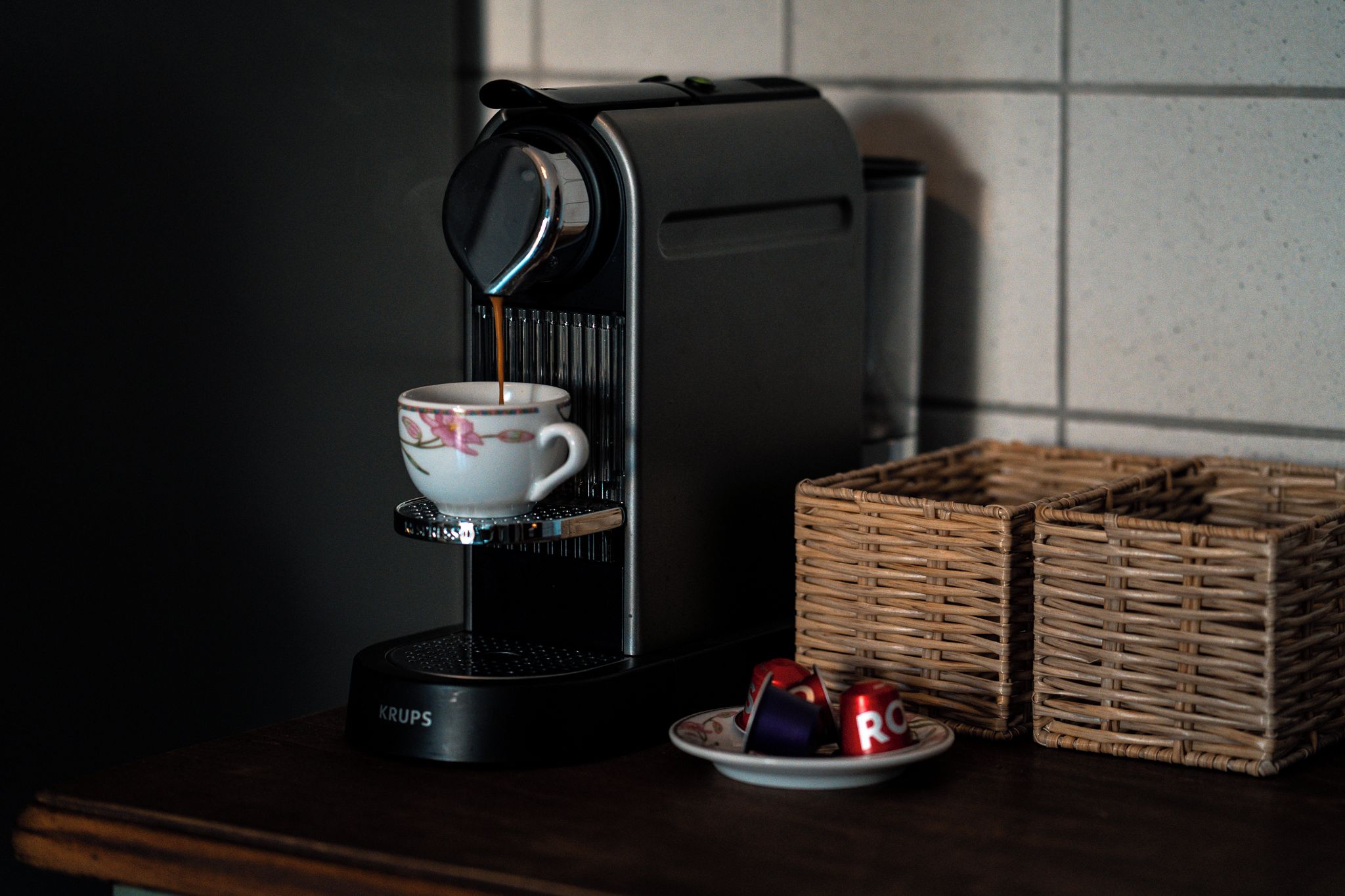 Una macchina da caffè con una tazza e un piattino, un piatto con una lattina e una tazza, e due cesti di vimini su un piano di lavoro scuro.
