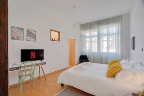 A hotel room with a bed, a desk with a TV, a chair, and a window with curtains.
