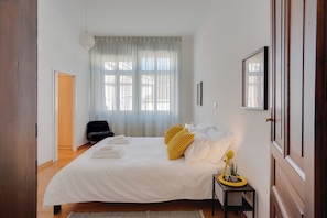 A bedroom with a bed, a bedside table with a lamp, a potted plant, and a window with sheer curtains. - Chromatica - di Fronte a Galleria Bomb (Gorizia)