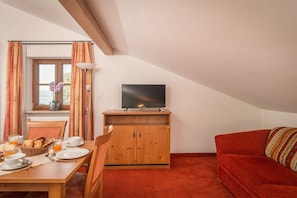 A cozy room with a dining table set for breakfast, a television on a wooden stand, and a red sofa. - Wiesholler Guest and Apartment House (Chieming)