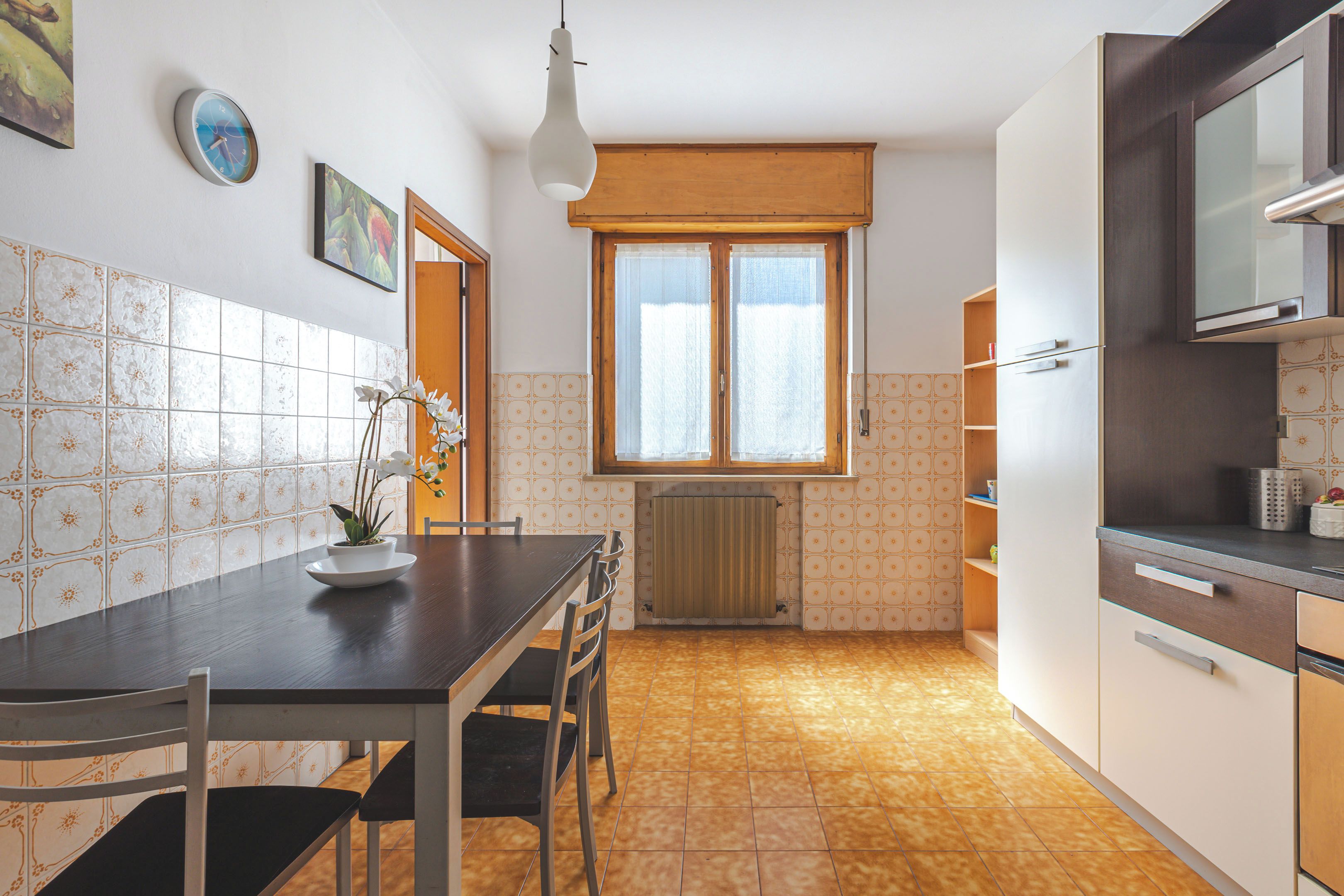 A compact kitchen with a dining table, chairs, a hanging light fixture, and a window with a wooden blind.