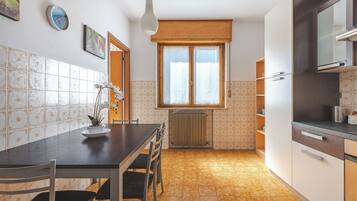 A compact kitchen with a dining table, chairs, a hanging light fixture, and a window with a wooden blind.
