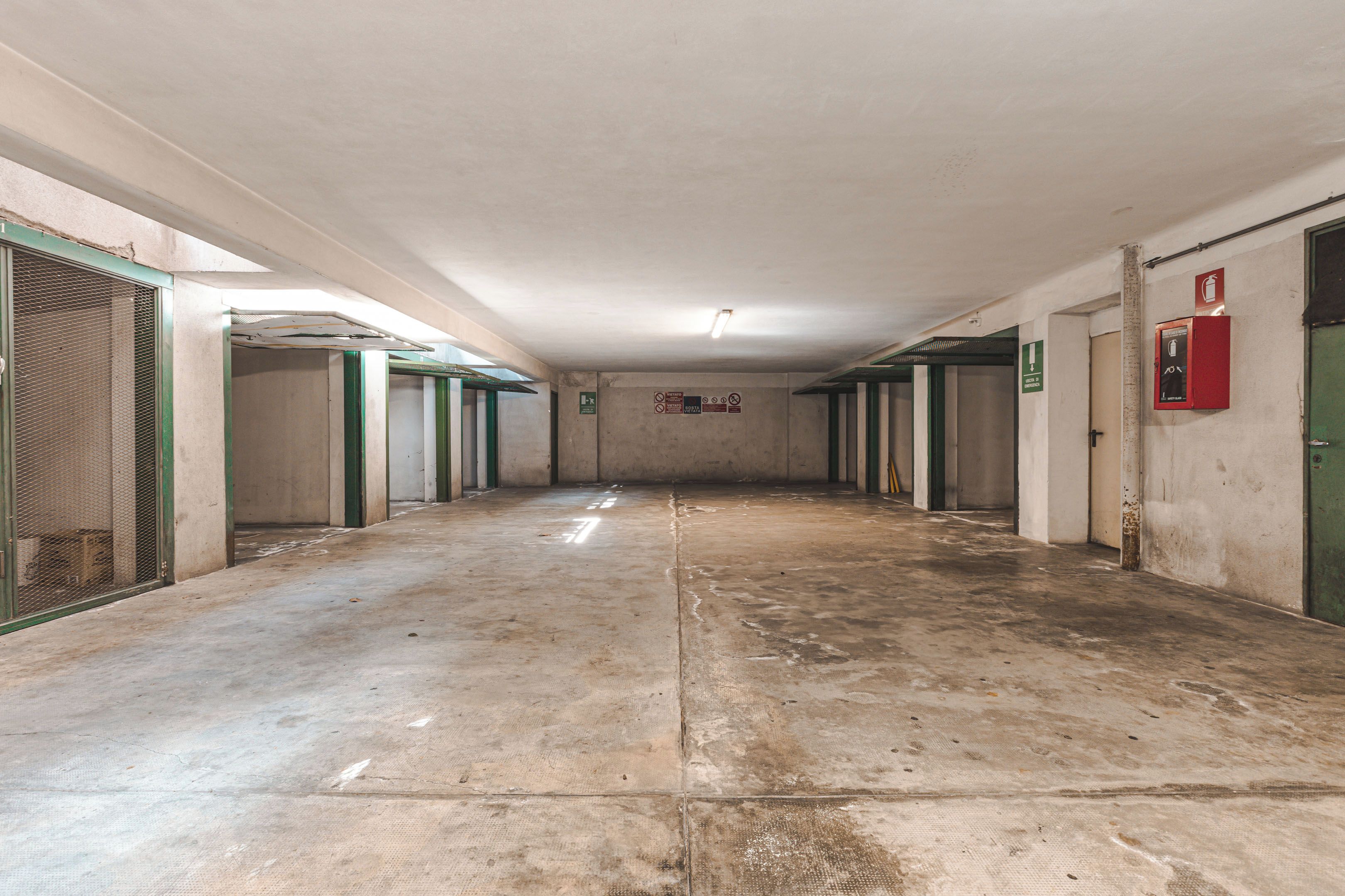 A long corridor with doors on both sides, some with green frames, and a red emergency equipment box on the right wall.