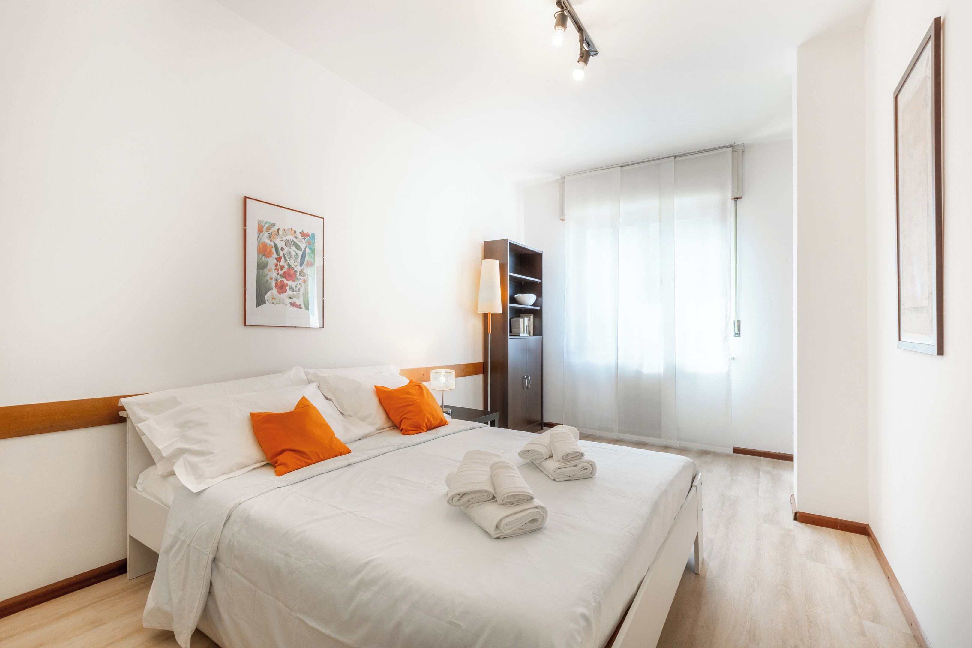 Habitación de hotel con cama, almohadas, mesita de noche, lámpara, armario y un cuadro en la pared.