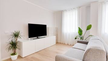 Een moderne woonkamer met een flatscreen televisie op een wit kastje, een grijze bank en twee potplanten.