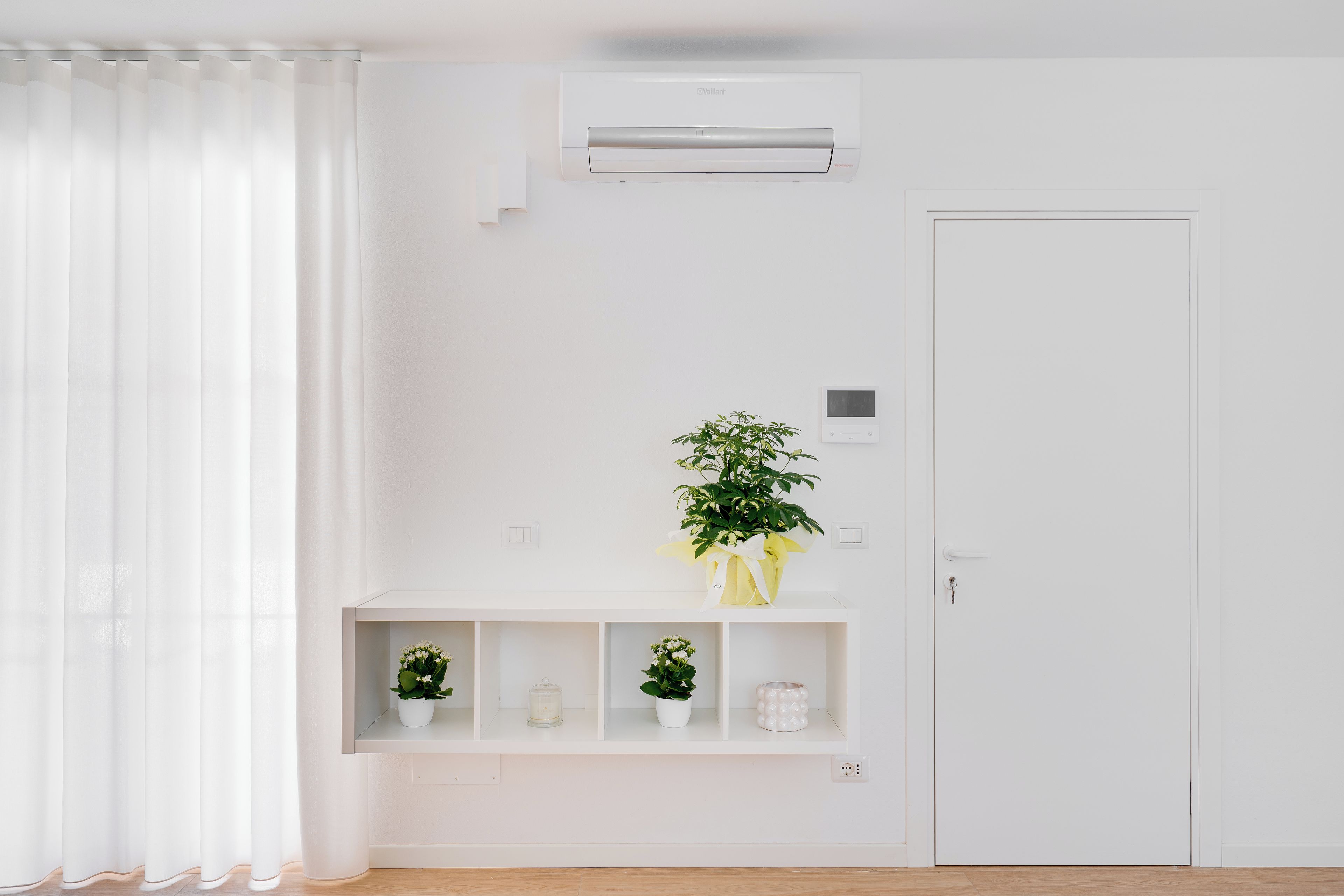 A minimalist room with a white door, an air conditioning unit, a shelf with potted plants, and a white curtain.