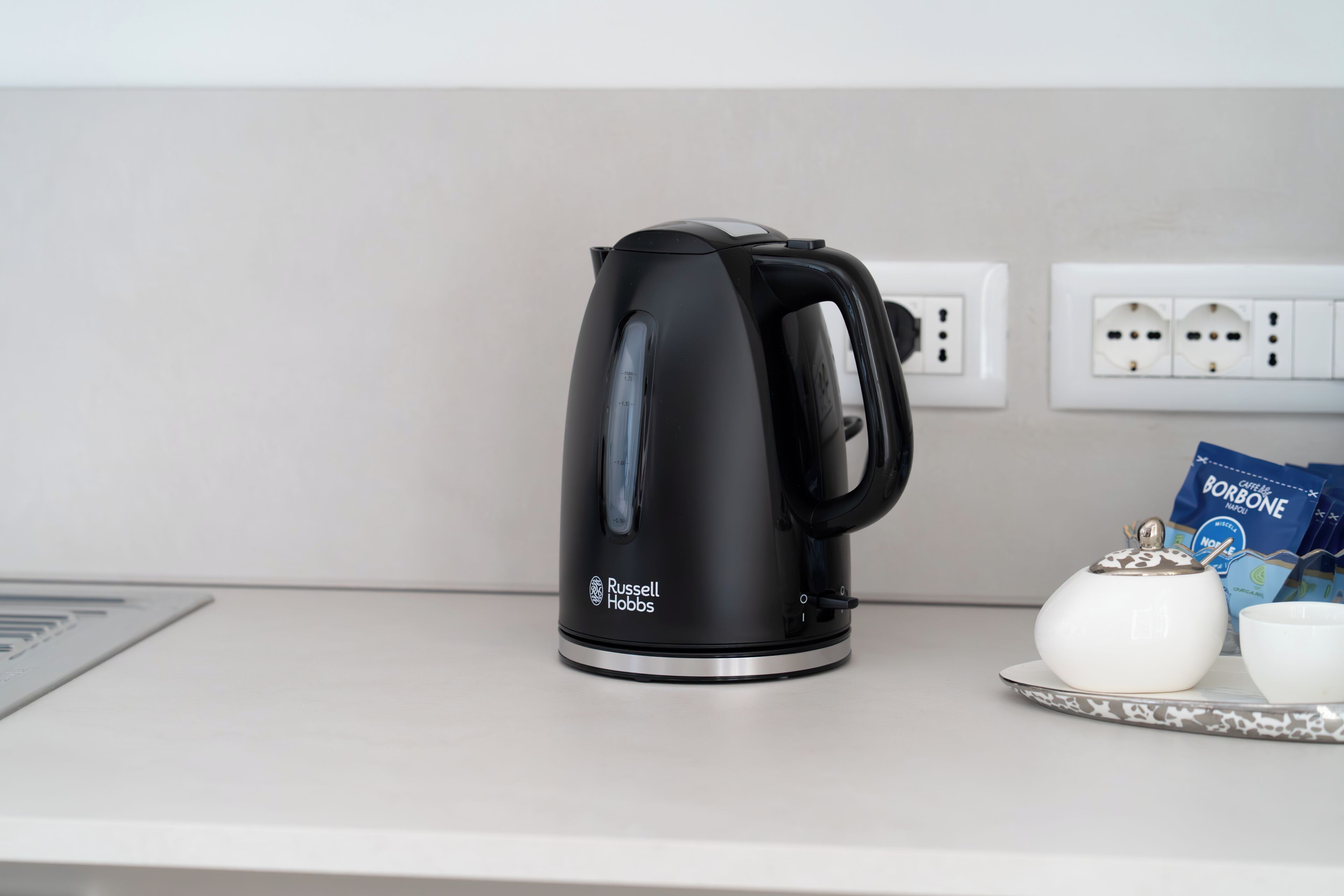 A black Russell Hobbs electric kettle on a kitchen counter with a white teapot and a blue packet of coffee.
