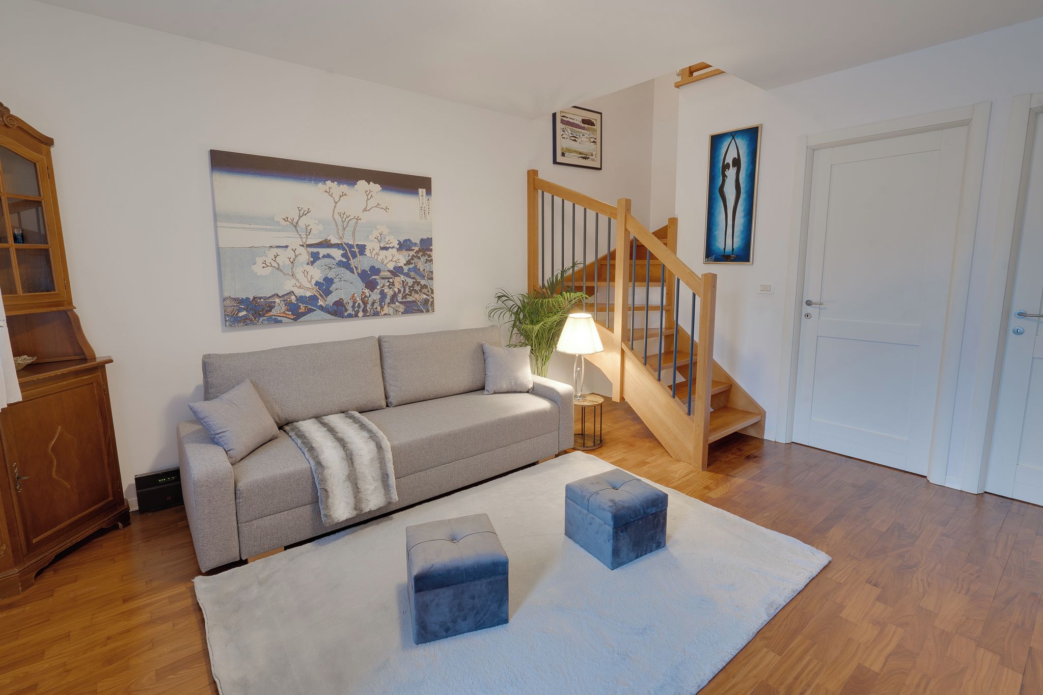 Un salon avec un canapé gris, un meuble en bois, un tableau, un escalier et une porte.