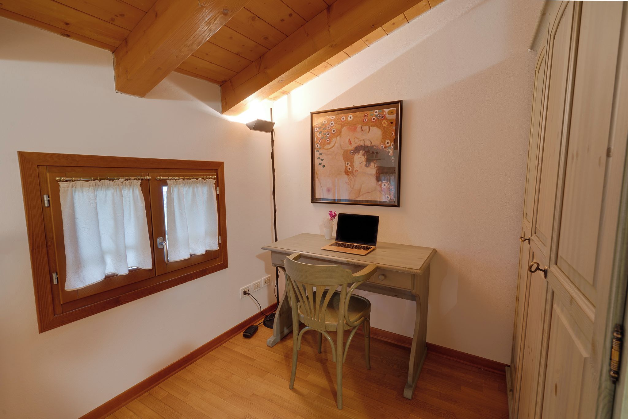 A room with a wooden desk, a chair, a laptop, a window with curtains, and a painting on the wall.