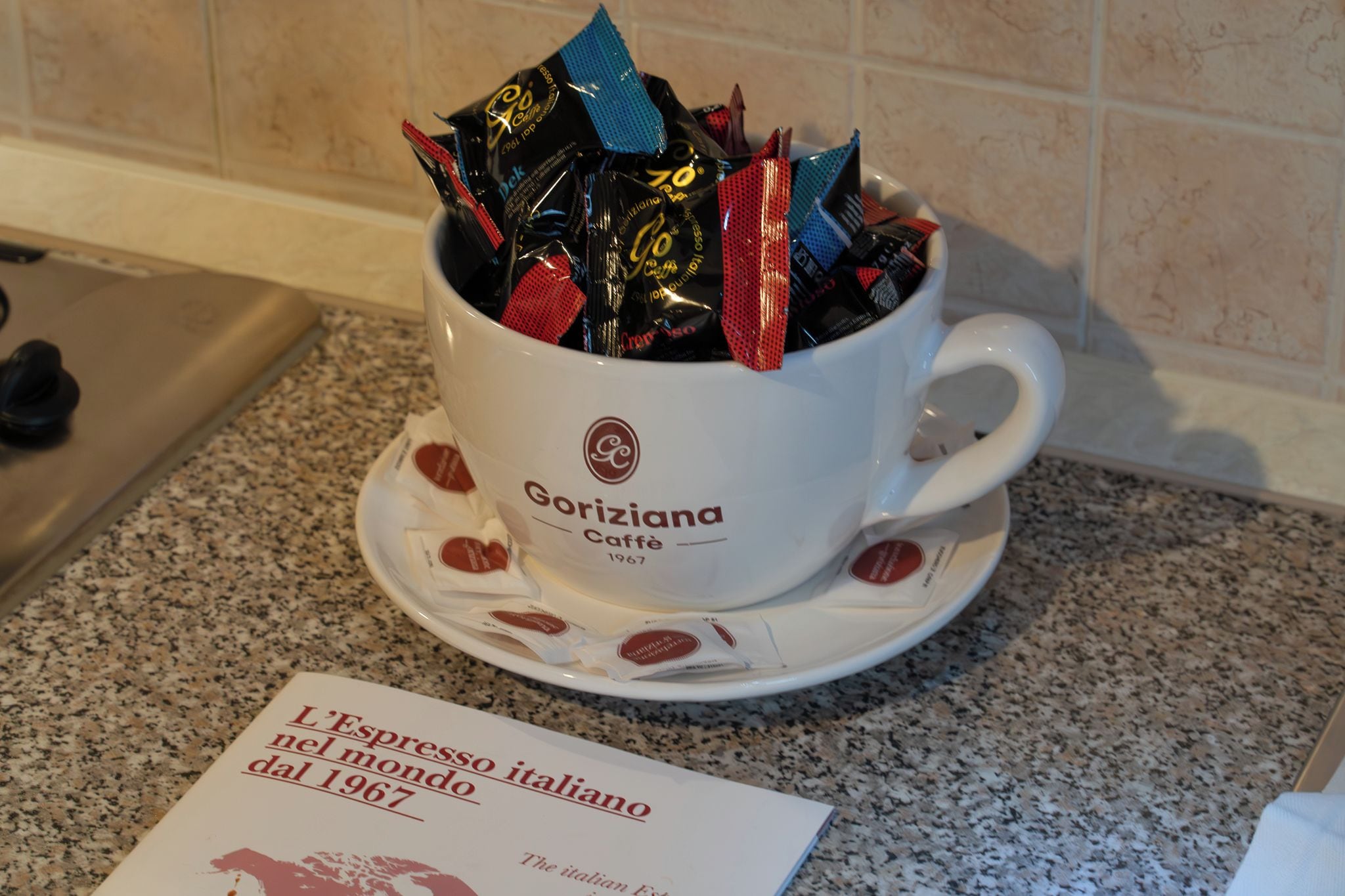 Une tasse blanche ornée d’un logo et d’un texte, remplie de différents sachets de café, posée sur un comptoir en granit.