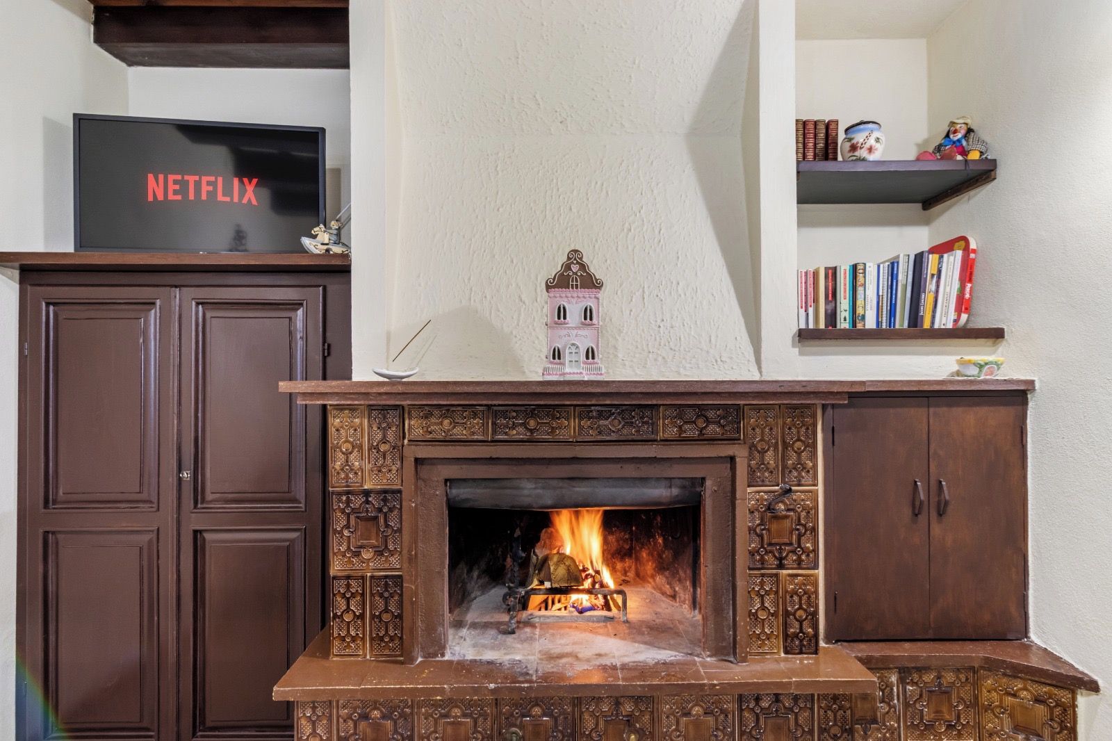 Una chimenea con diseños intrincados, un televisor mostrando 'NETFLIX' y una estantería con libros y objetos decorativos.