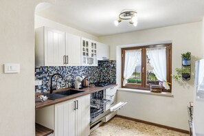 Een kleine keuken met witte kasten, een betegelde spatwand, een raam met gordijnen en een inbouw vaatwasser.