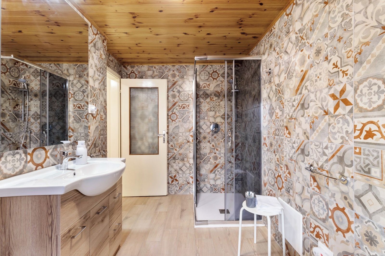 Ein Badezimmer mit Holzboden, gemusterten Fliesen, einer Duschkabine aus Glas, einem weißen Waschbecken und einem kleinen weißen Tisch.