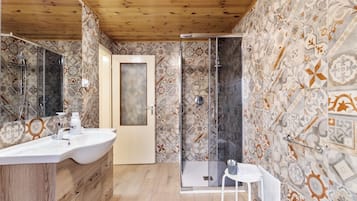 Ein Badezimmer mit Holzboden, gemusterten Fliesen, einer Duschkabine aus Glas, einem weißen Waschbecken und einem kleinen weißen Tisch.
