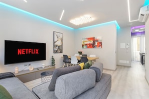 Una sala moderna con un televisor de pantalla plana mostrando 'NETFLIX', un sofá, una mesa de centro y una zona de comedor con sillas y una mesa.