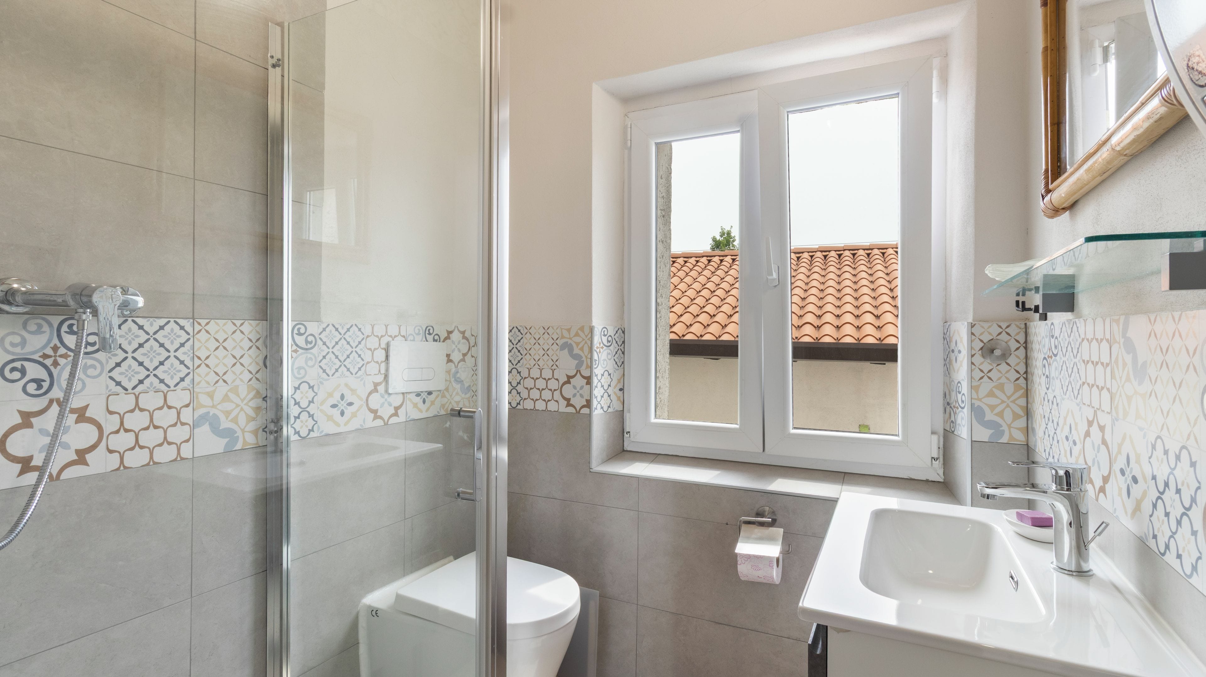 Baño con ducha, inodoro y lavamanos, con azulejos estampados y una ventana.