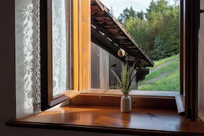 A window with a wooden sill, a white vase with a plant, and a view of a wooden structure and greenery outside.