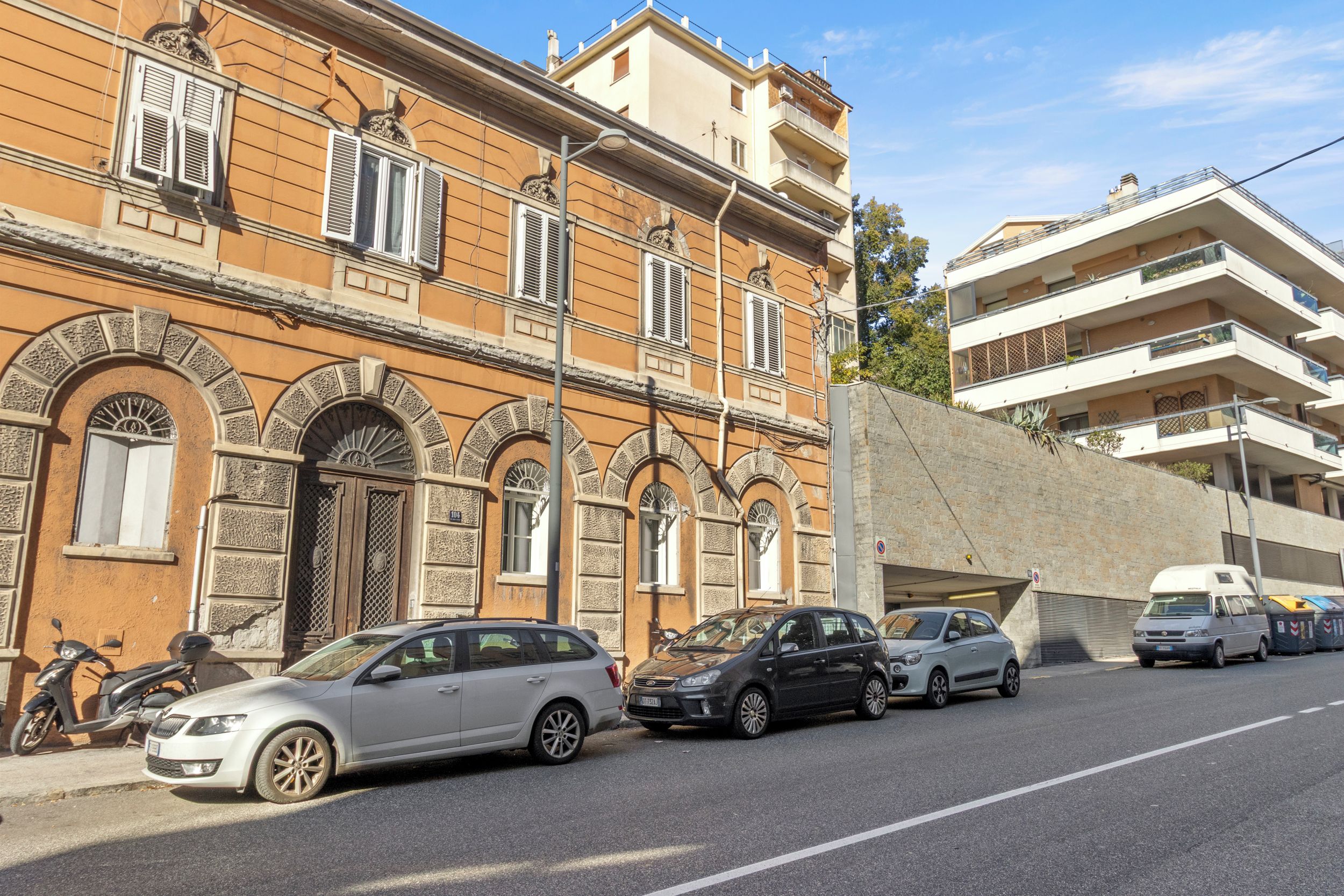 Eine Straße mit parkenden Autos und einem Gebäude mit rundbogenförmigen Fenstern und einer Steinaußenwand.