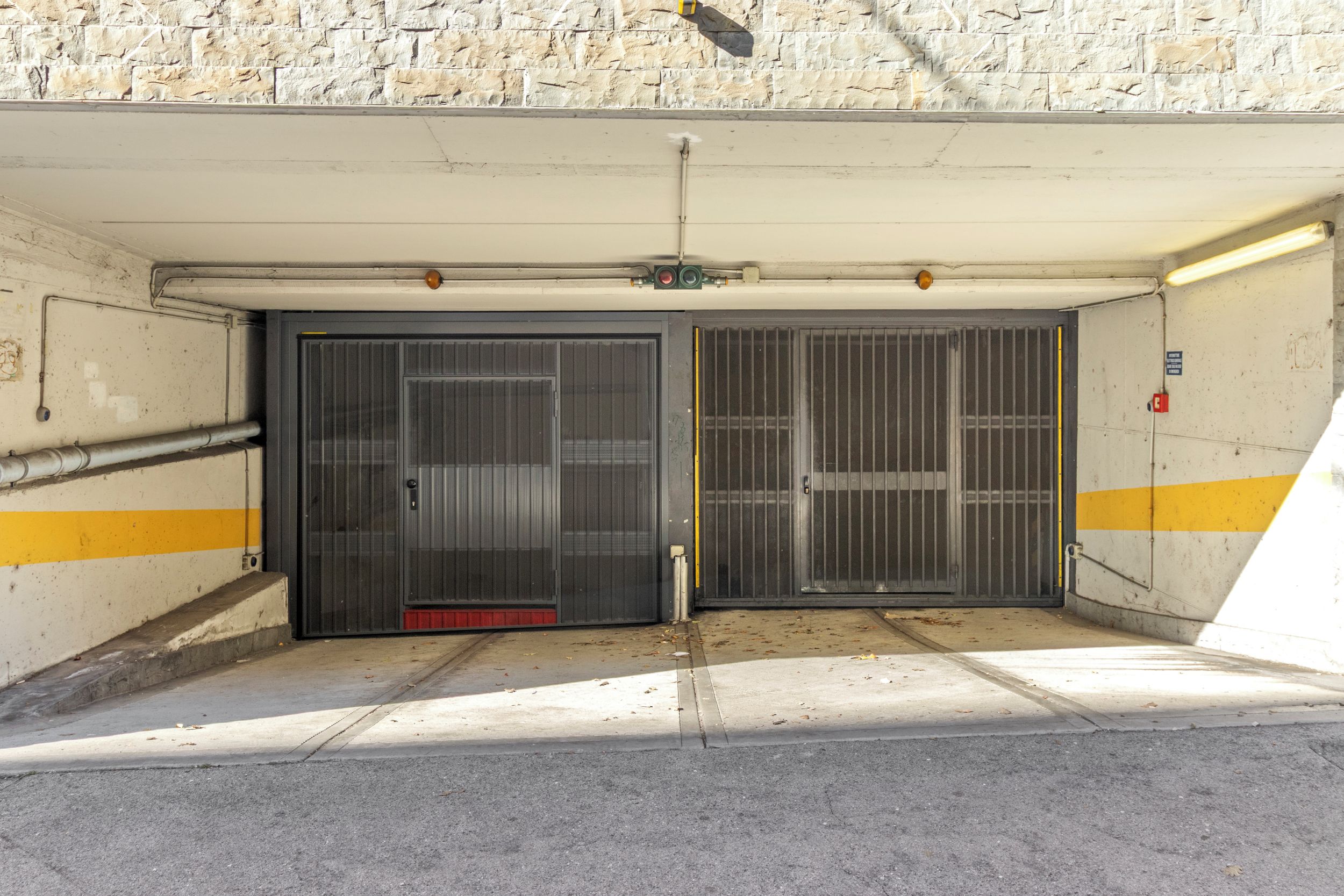 L’entrée d’un parking, avec deux portes métalliques, un sol en béton et un mur en pierre.