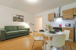 A compact living space with a green sofa, a dining table with chairs, a kitchen area with wooden cabinets, and a painting on the wall. - New! Tocai in via Rastello per 4 pp (Gorizia)