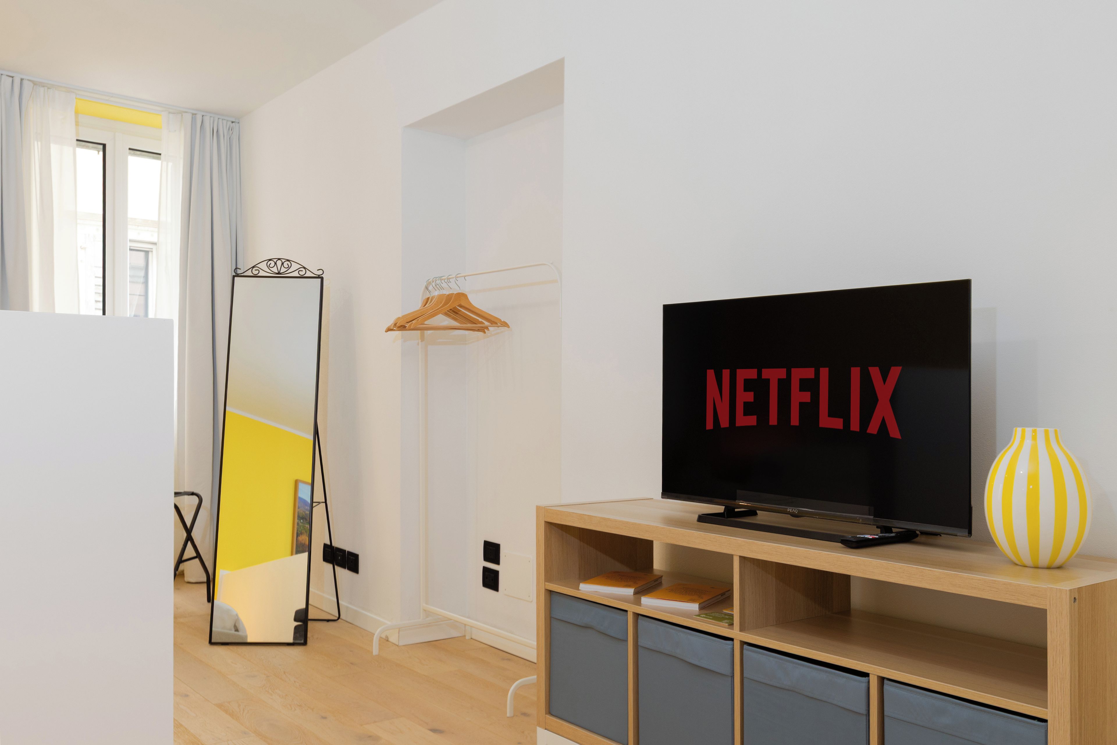 Un soggiorno moderno con una TV a schermo piatto che mostra il logo di Netflix, un mobile TV in legno, uno specchio e un vaso giallo.