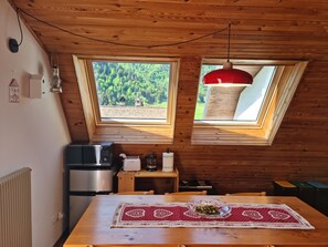 Een gezellige keuken met een houten plafond, een tafel met een kantkleed en uitzicht op een bos door twee ramen.