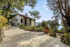 A stone pathway leading to a white building with a green roof, surrounded by trees and a view of the sea.