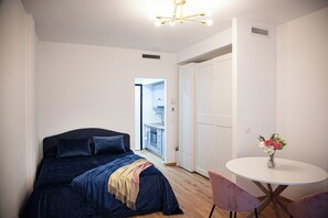 Quarto com cama, mesa de cabeceira com um vaso de flores, mesa redonda com cadeiras e vista para uma varanda.