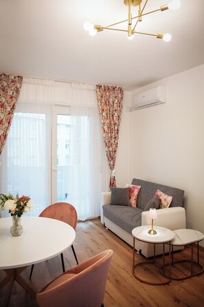 Uma sala de estar moderna com sofá, uma mesa redonda e uma cadeira. Há uma janela com cortinas leves e uma cortina estampada com flores.