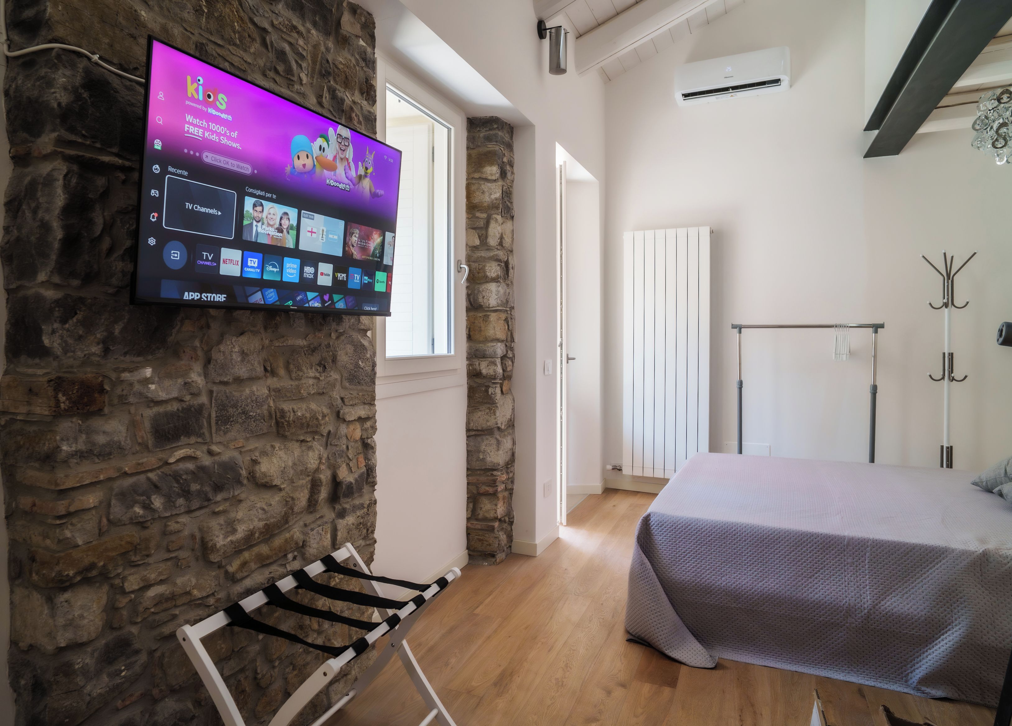 Una camera moderna con parete in pietra, una TV a schermo piatto che mostra un canale per bambini, un letto con copriletto grigio e pavimento in legno.