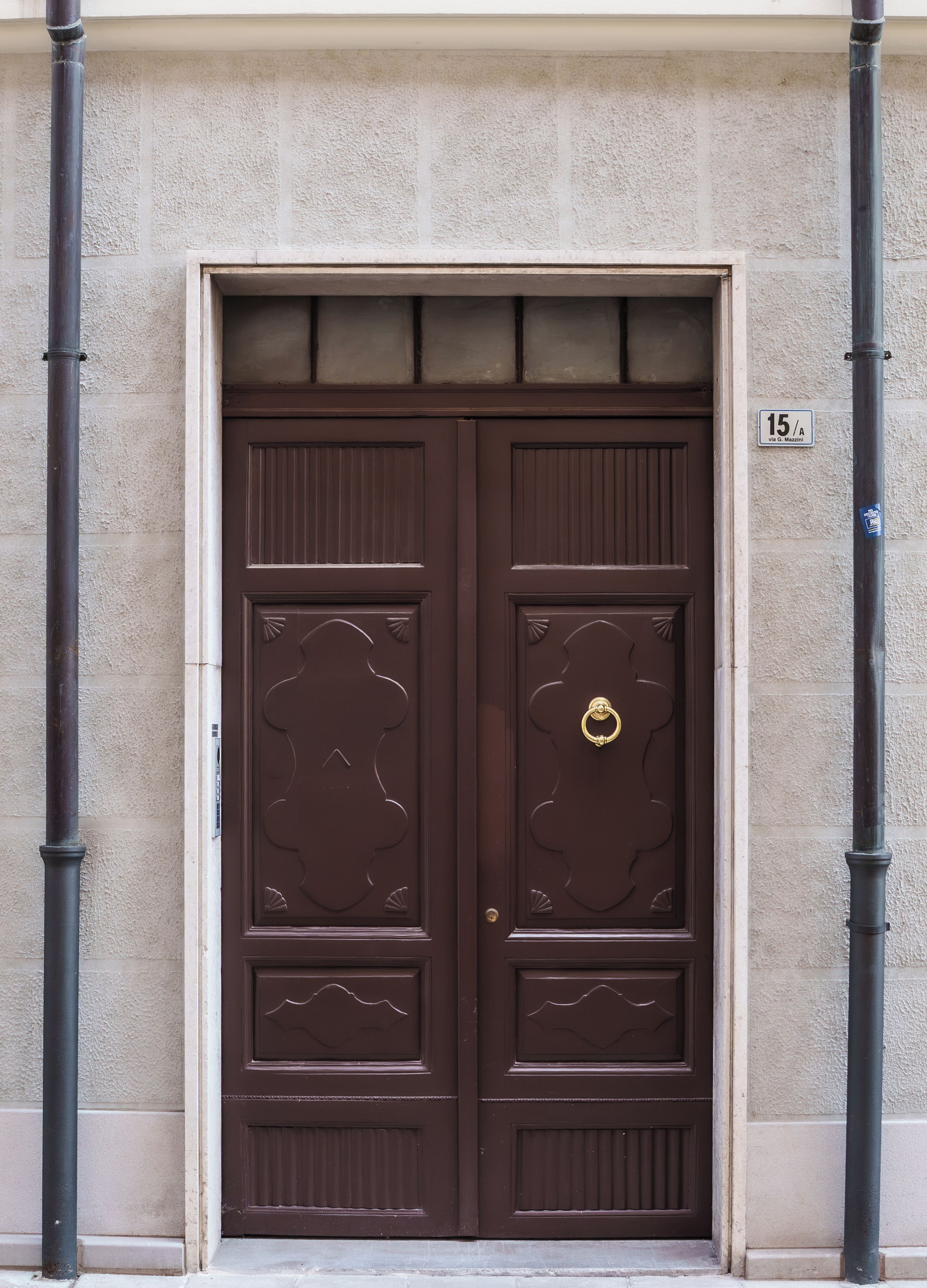 Una puerta doble con un aldabón de latón, integrada en una pared beige con textura, con una placa de número '15/A' a la derecha.