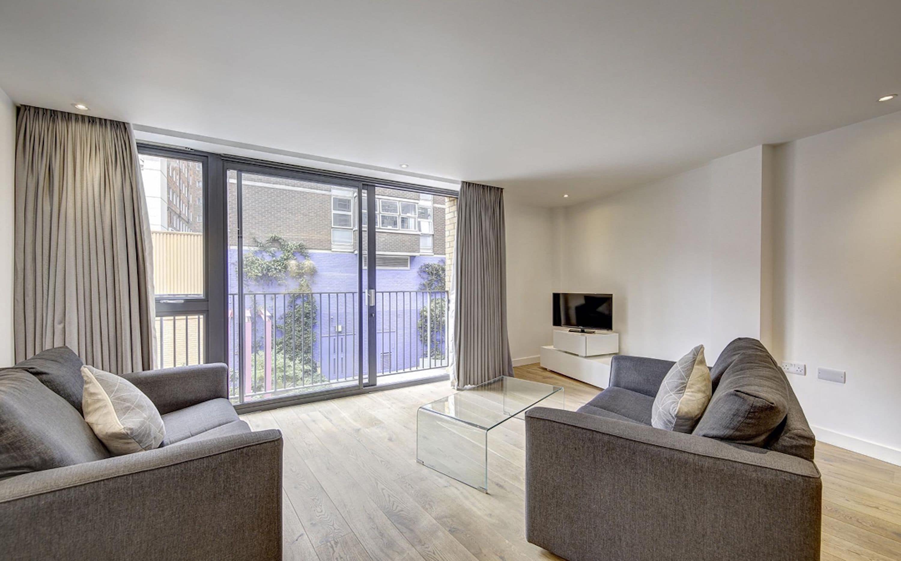 A modern living room with a glass coffee table, two grey sofas, a flat-screen TV, and a large sliding glass door leading to a balcony.