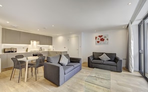 Ein modernes Wohnzimmer mit einem grauen Sofa, einem Essbereich mit einem Glastisch und einer Küche im Hintergrund.