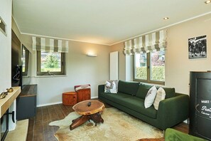 Een woonkamer met een groene bank, een ronde houten salontafel, een klein oranje voetenbankje en een vachtkleed.