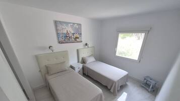 A bedroom with two single beds, a nightstand, a small table, a window, and a painting on the wall.