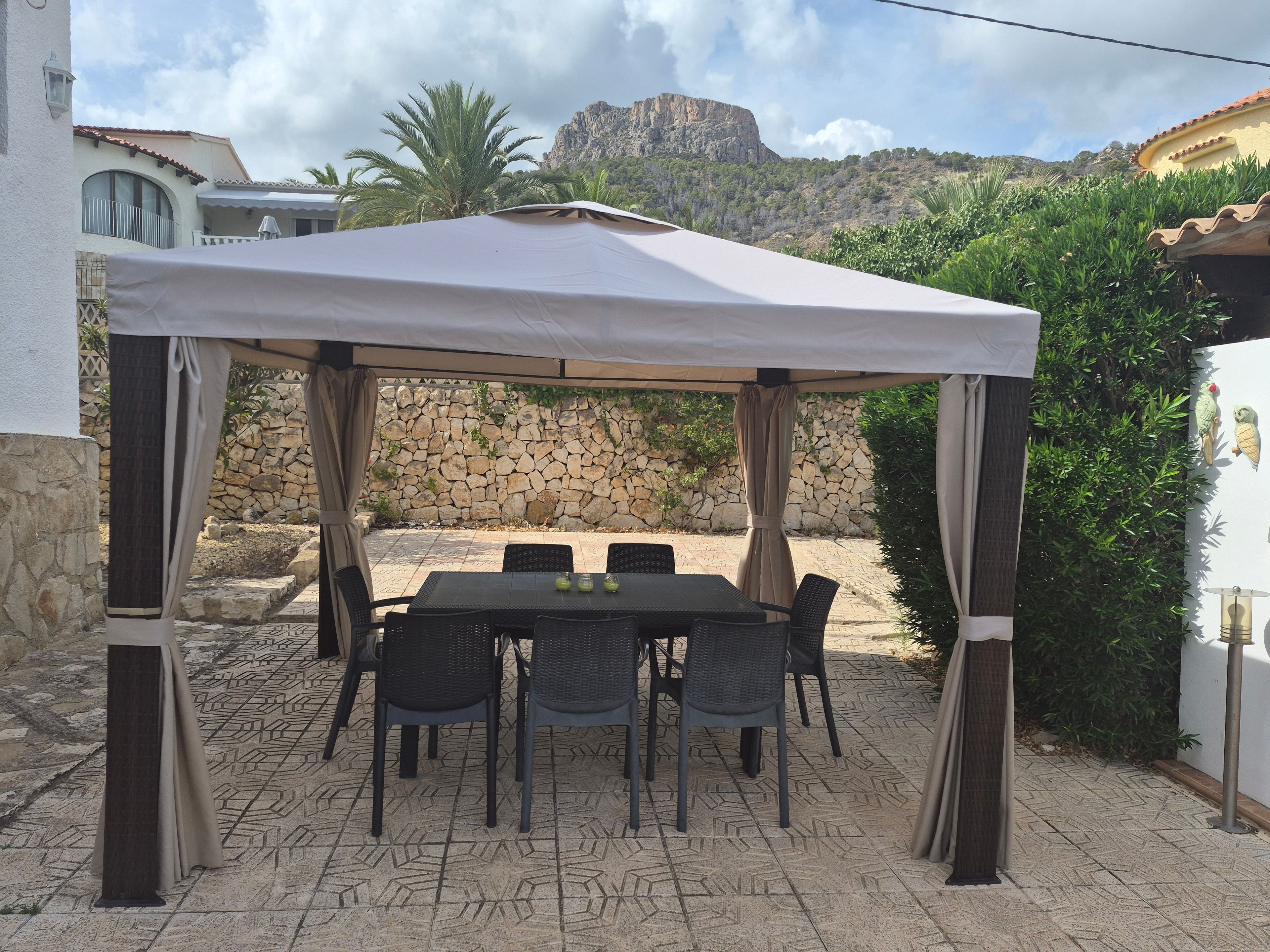 An outdoor patio with a table and chairs under a canopy, surrounded by stone walls and vegetation.