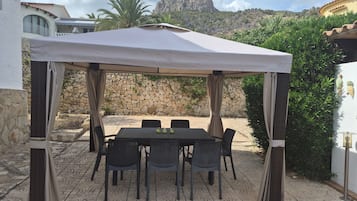 An outdoor patio with a table and chairs under a canopy, surrounded by stone walls and vegetation.