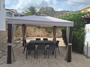 Een terras buiten met een tafel en stoelen onder een zonnescherm, omringd door stenen muren en begroeiing.