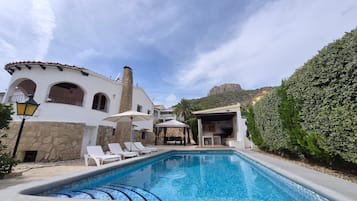 A swimming pool with lounge chairs, a stone building with arches, and a view of a mountain.