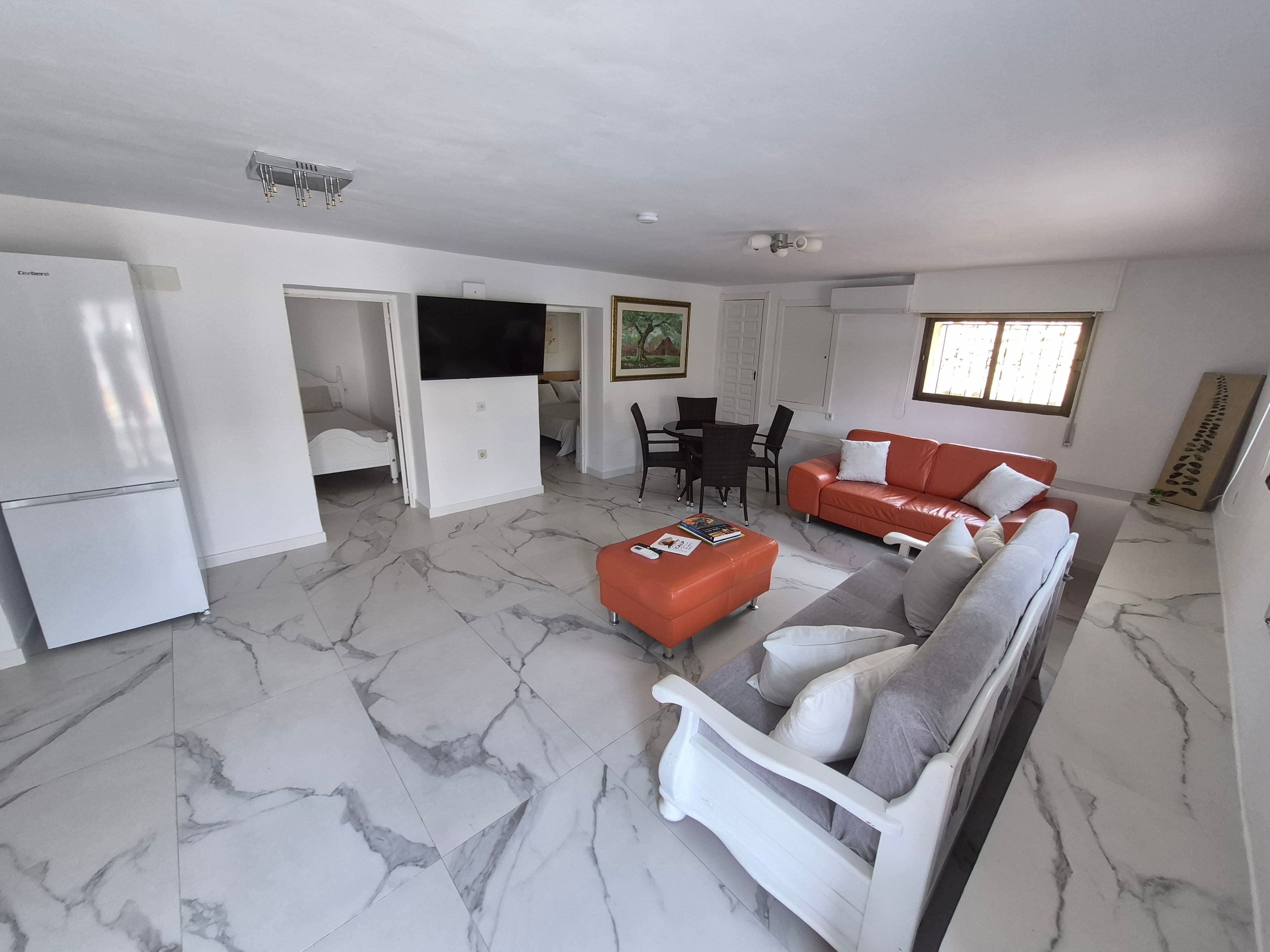 Een ruime woonkamer met een witte bank, een rode bank, een eettafel met stoelen, en een keuken met een koelkast.